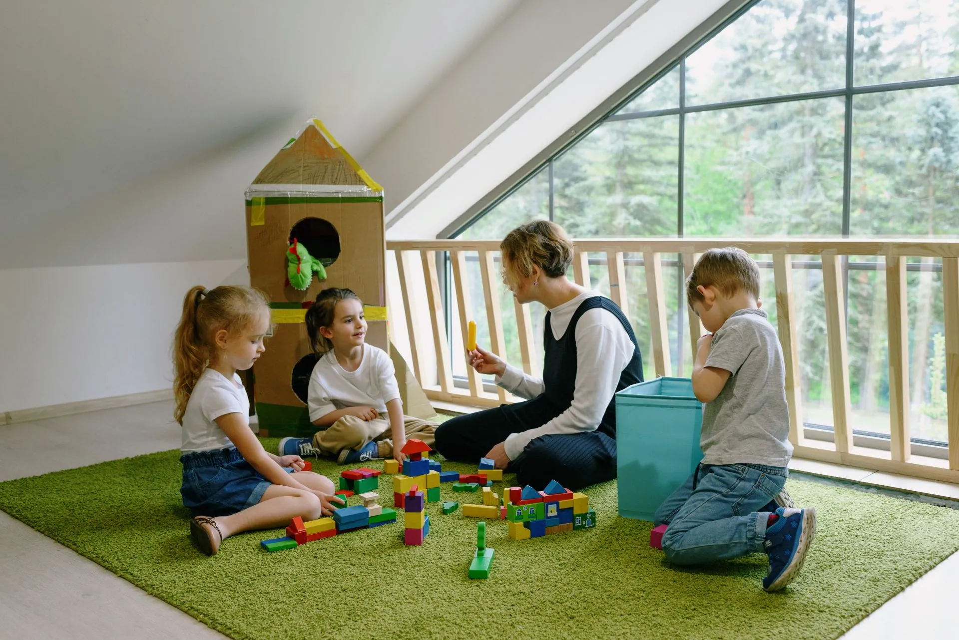 Eine Erzieherin spielt mit drei Kindern Bauklötze auf einem grünen Teppich im hellen Spielzimmer